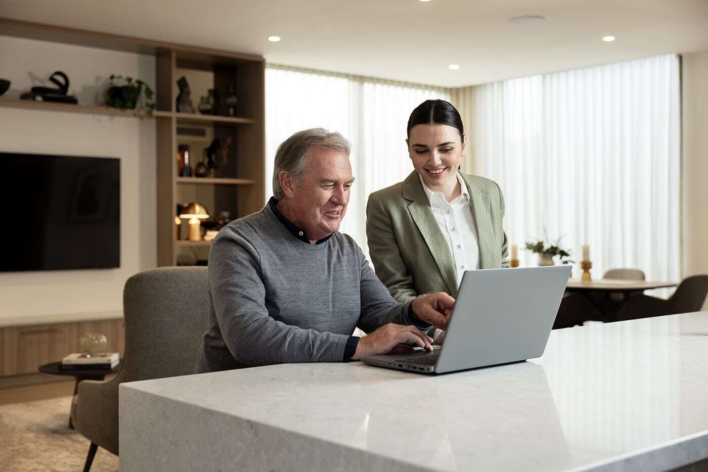 Older man using a laptop at a kitchen bench with a younger woman standing beside him, both smiling and looking at the screen.