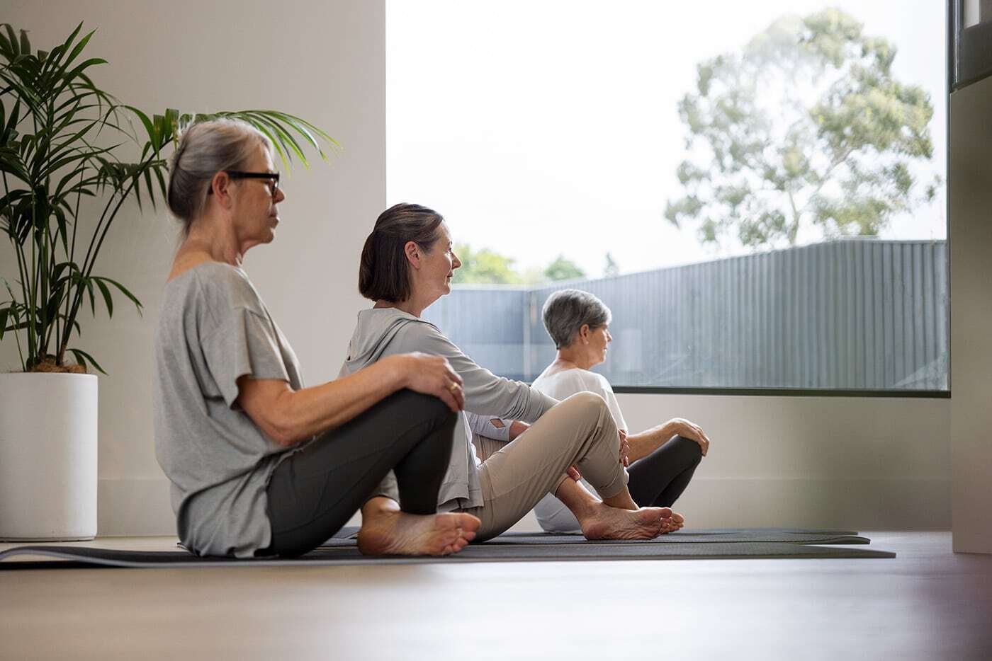 Three older women seated on yoga mats in a light-filled room, stretching and looking out a large window.