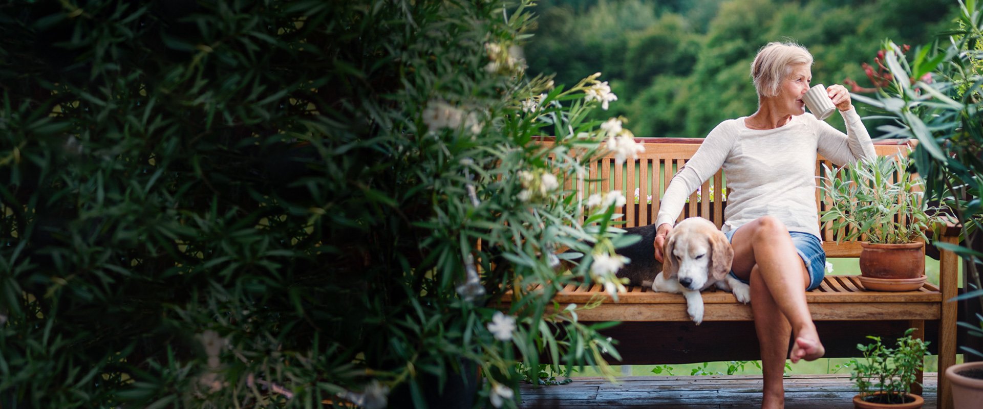 Woman sits on a wooden garden bench surrounded by plants, sipping from a mug while petting a relaxed beagle lying beside her.