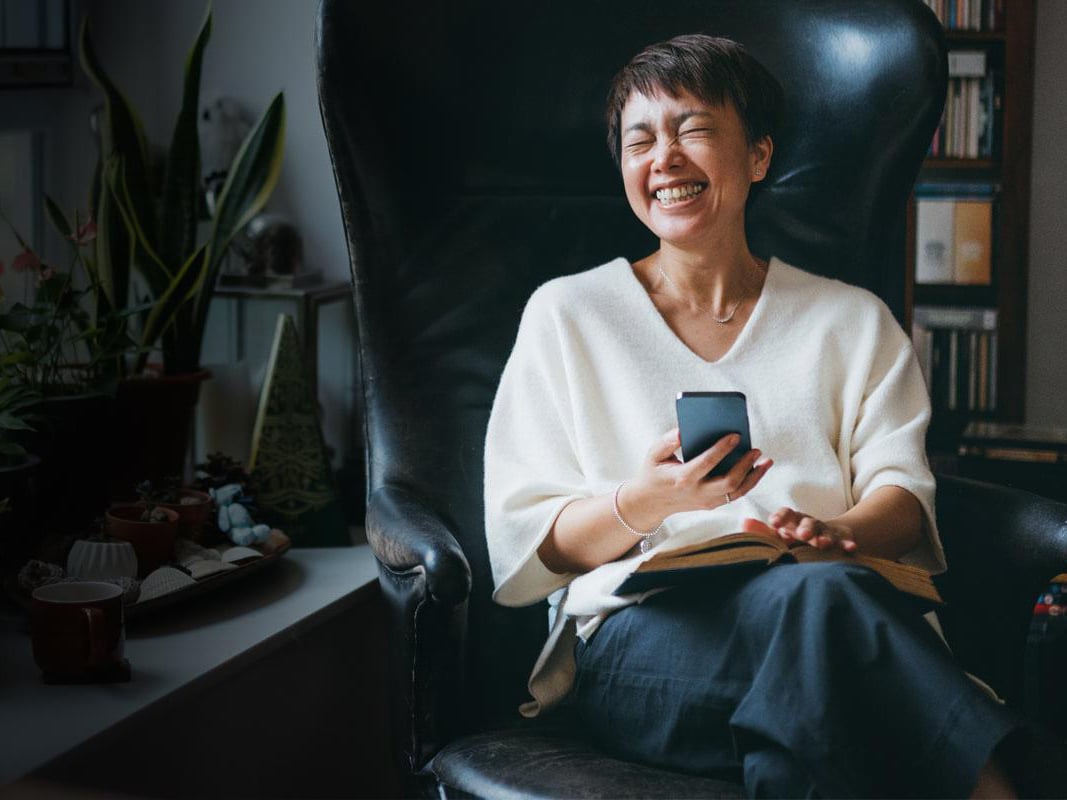 Woman sits on chair laughing with phone in hand